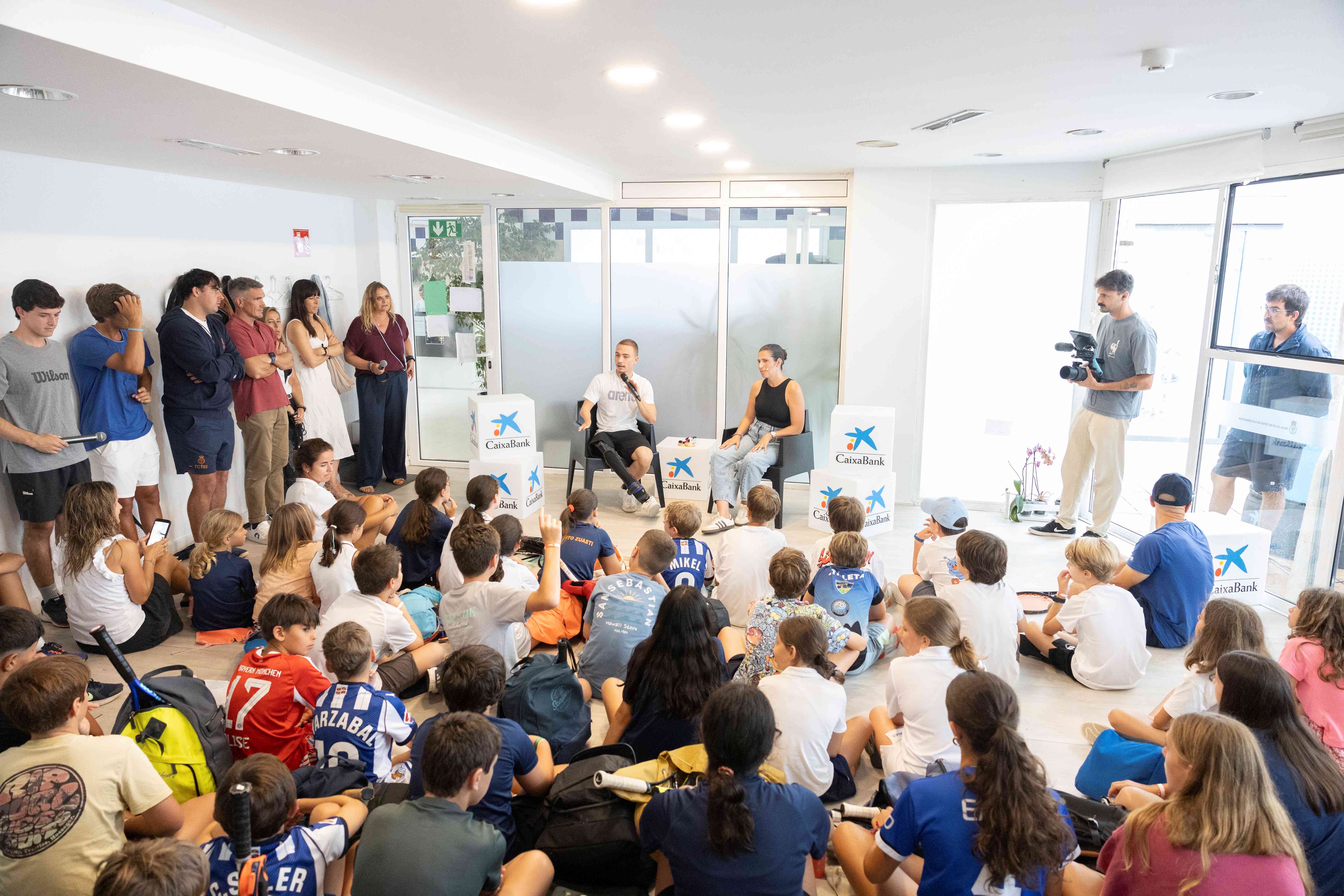 Activación CaixaBank Empower Kids en Tenis San Sebastián: decenas de niños y niñas sentados en el suelo escuchan a un deportista paralímpico mientras Carmen de Zarata conduce el encuentro, rodeados de cubos con el logo de CaixaBank.
