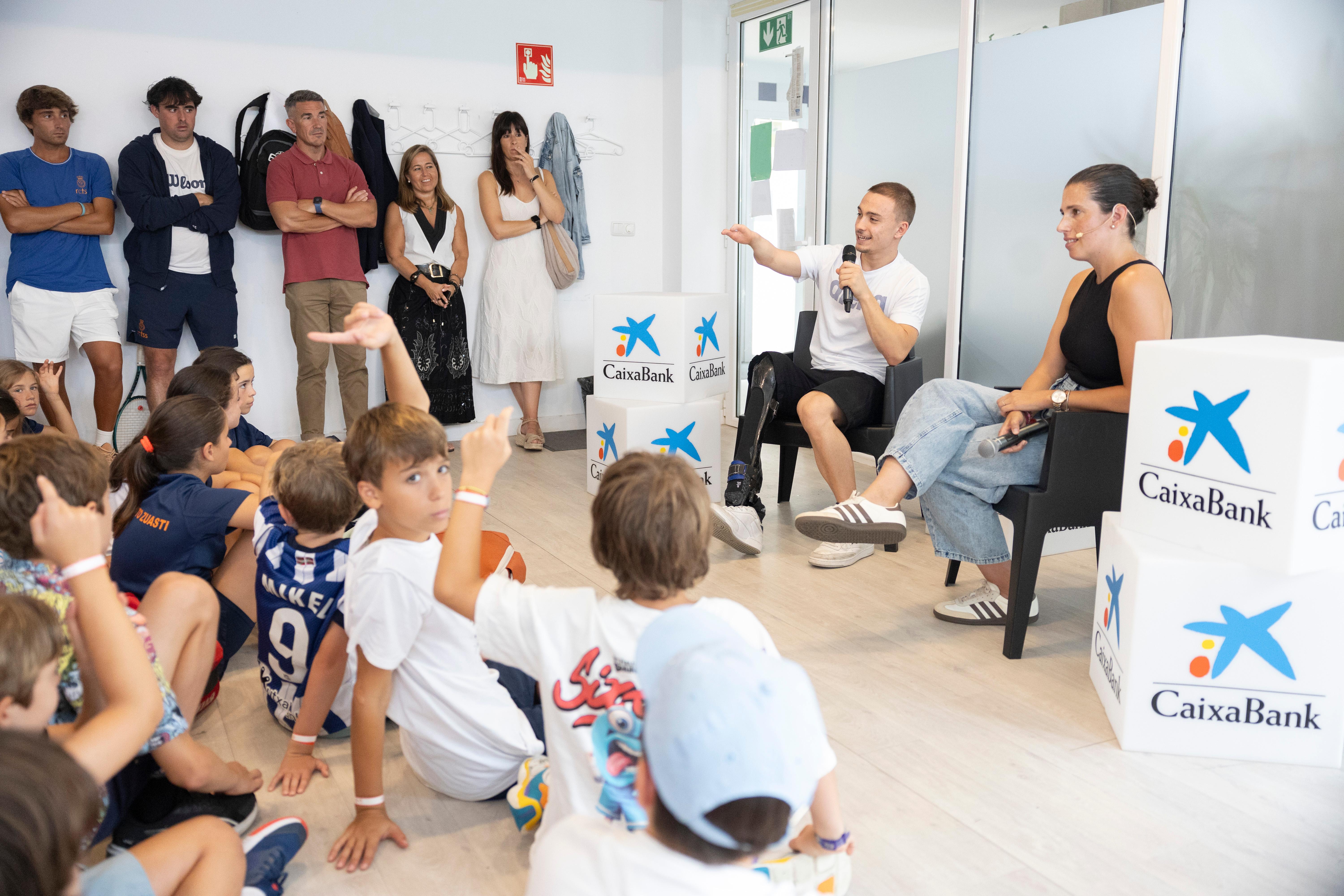 Carmen de Zarata moderando la activación CaixaBank Empower Kids en San Sebastián, en conversación con un deportista paralímpico ante un grupo de niños y niñas sentados en el suelo, entre cubos con el logo de CaixaBank.
