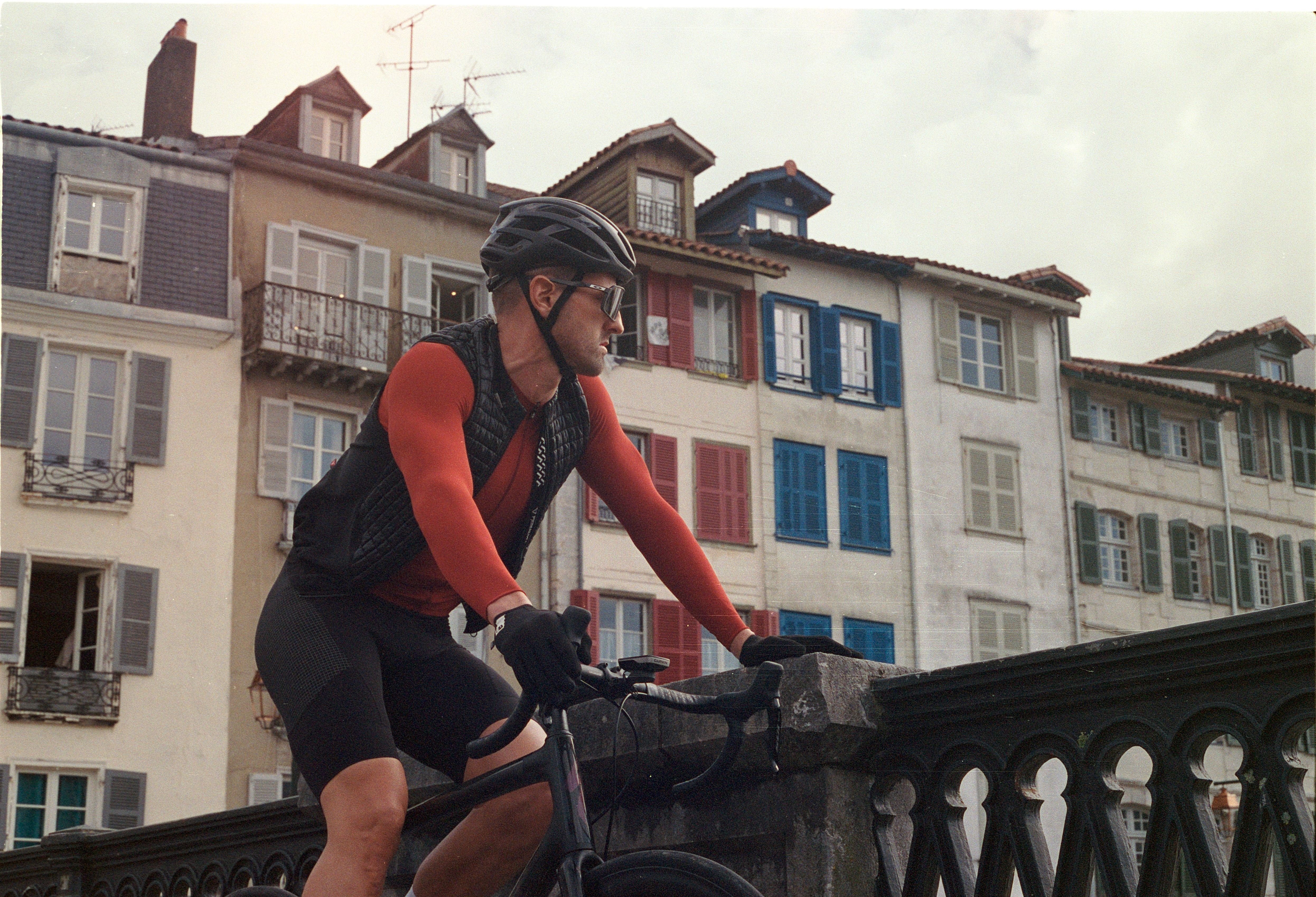 Ciclista de la campaña Etxeondo apoyado sobre el pretil de un puente, con fachadas de contraventanas de colores al fondo.