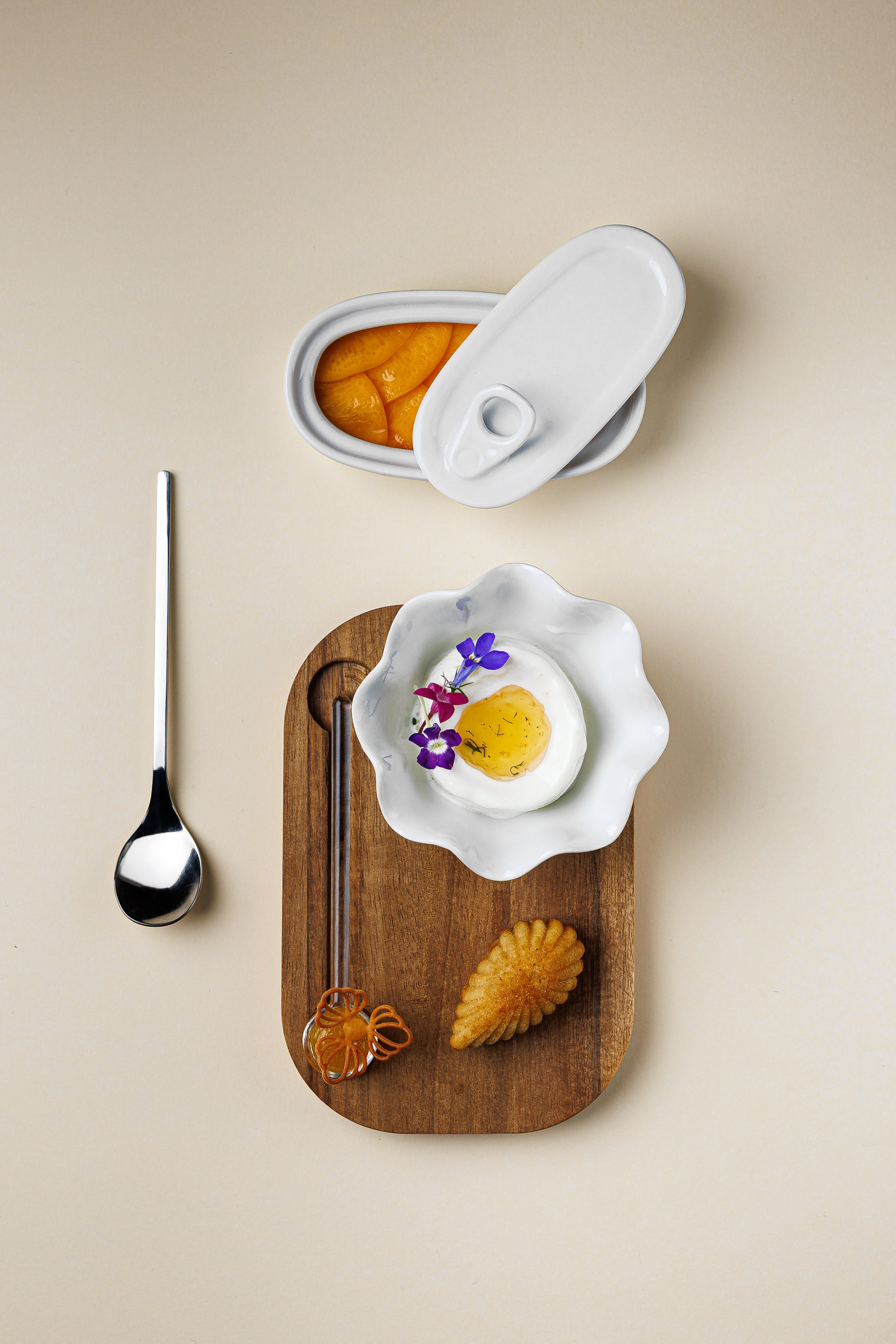 Postre de alta cocina de INA visto en cenital: una lata de conserva cerámica con gajos de cítrico, un cuenco con forma de flor que sostiene un postre que imita un huevo frito coronado con violetas comestibles, y una madalena con forma de concha sobre una tabla de madera.