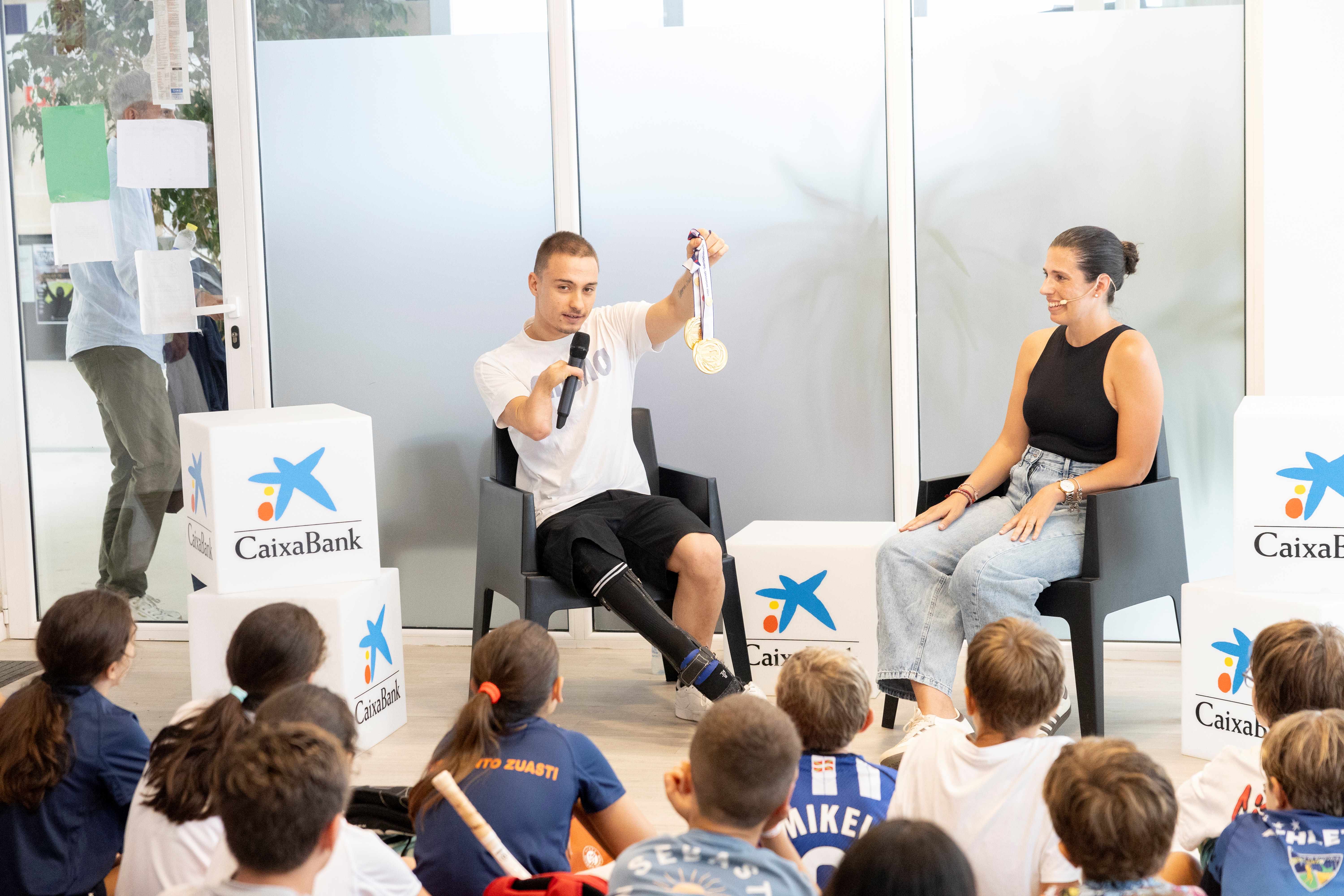 Un deportista paralímpico, sentado y con micrófono, muestra sus medallas de oro mientras Carmen, sentada frente a él, modera el encuentro ante un grupo de niños sentados en el suelo, entre cubos con el logo de CaixaBank.