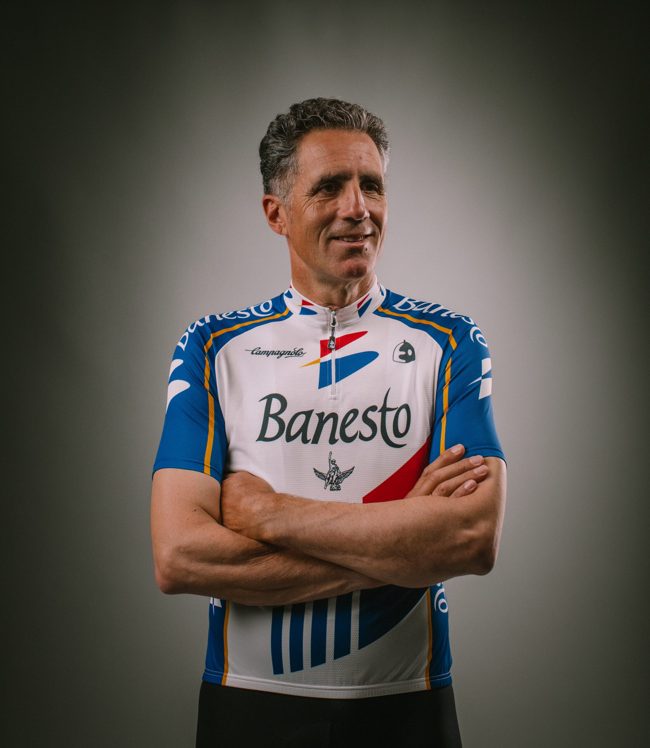 Miguel Induráin, con el maillot blanco y azul de Banesto y los brazos cruzados, posa sonriente sobre un fondo gris de estudio.
