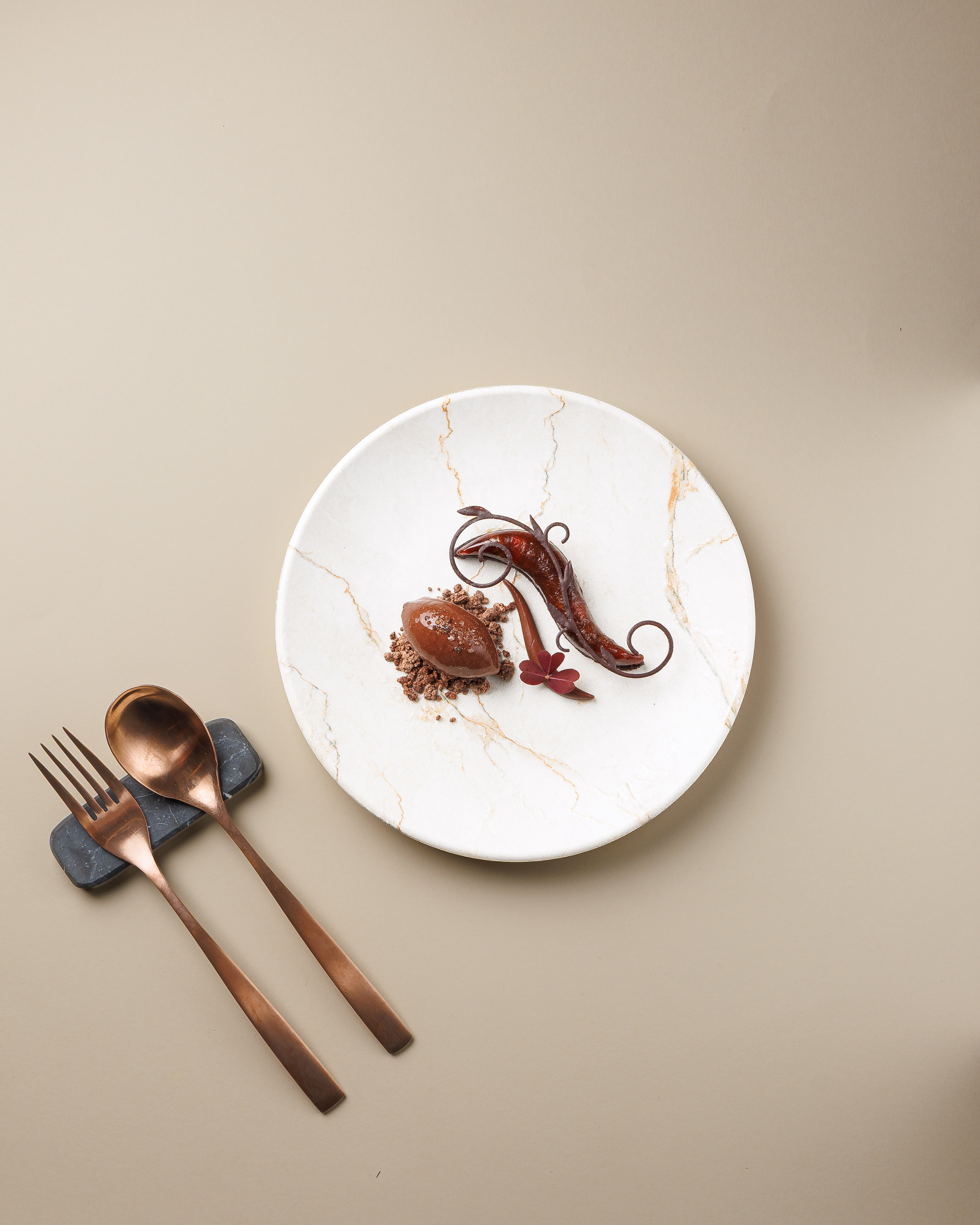 Postre de alta cocina emplatado sobre un plato de mármol claro, con una quenelle de chocolate y decoración, junto a cubiertos de cobre sobre fondo neutro.
